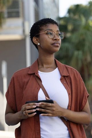 Young african american woman holding smartphone on urban street wearing glasses rust shirt