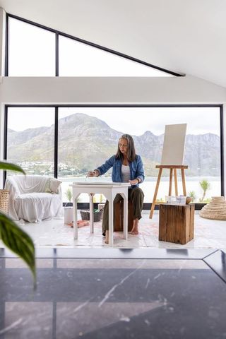 Senior woman painting in bright lakefront studio with mountain view, peaceful creativity