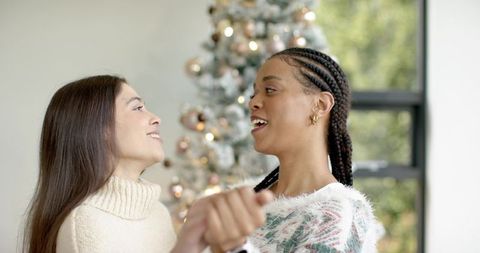 Diverse friends holding hands and swaying by christmas tree in cozy knit sweaters