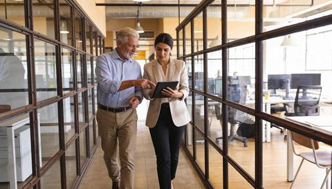 Senior mentor and young woman reviewing tablet while walking through open-plan office