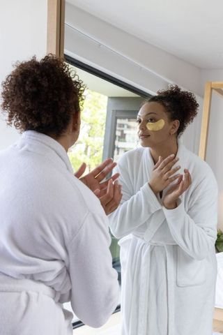 African american woman applying gold eye patches in white robe morning skincare ritual