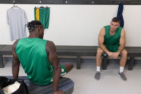 Male teammates sitting and talking in locker room wearing green jerseys preparing for game