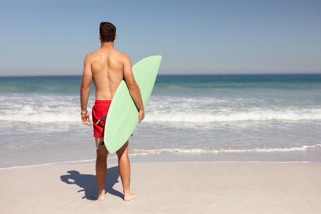Rear view of shirtless Caucasian man with surfboard standing on beach in the sunshine