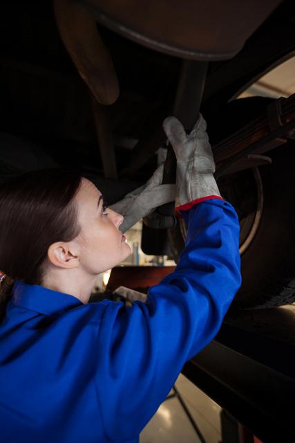 This image shows a female mechanic in a blue uniform and gloves, performing maintenance under a vehicle in a repair garage. It is ideal for use in content related to automotive services, gender equality in skilled trades, technical education, and professional labor advertisements.
