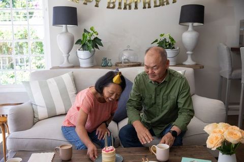 Asian couple celebrating intimate birthday at home with lit cake, roses, cozy living room