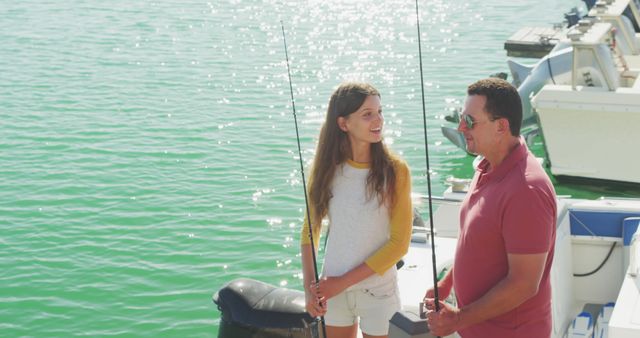 Happy caucasian teenage daughter and father talking on jetty with fishing rods on a sunny day. Leisure, free time, hobbies, family, travel and vacations.