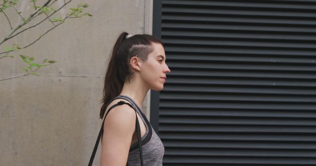 A young woman with a hearing aid walks confidently along an urban street. She is wearing fitness attire and has her dark hair pulled back in a ponytail. Ideal for use in materials related to fitness, hearing aids, independence, and empowerment.