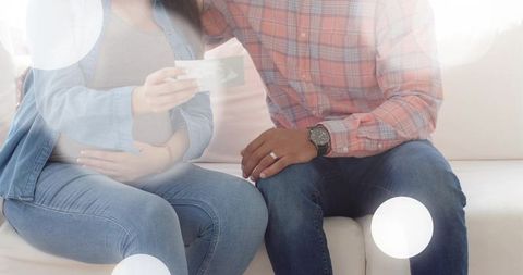 Expectant couple holding ultrasound, cradling pregnant belly on backlit living room sofa