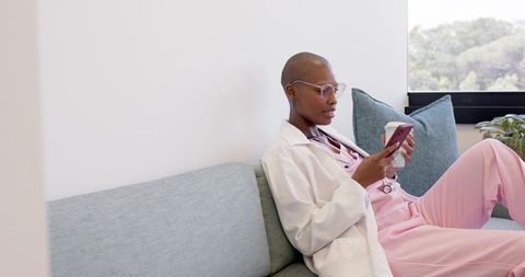 Relaxing clinician in white coat and pink scrubs checking smartphone on modern sofa holding coffee