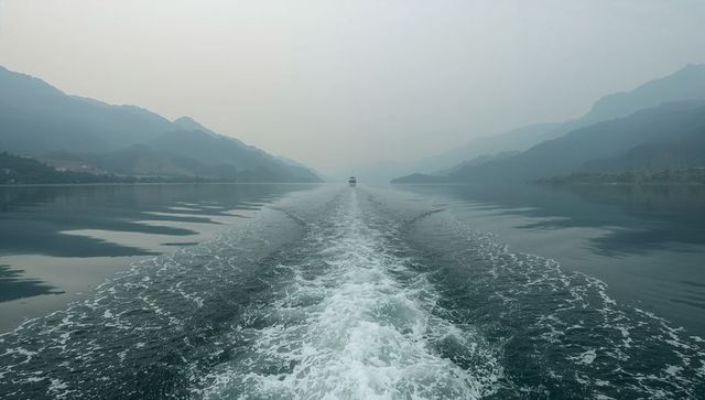 Boat wake cutting through misty alpine lake toward distant ferry and mountain ridges