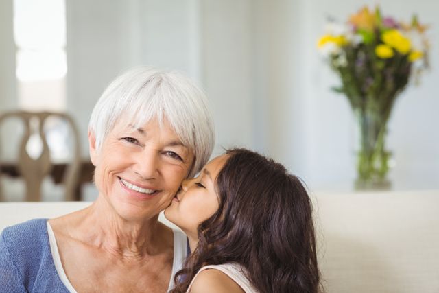 This image captures a heartwarming moment between a grandmother and her granddaughter in a cozy living room. The grandmother is smiling warmly as the young girl kisses her on the cheek, showcasing a strong family bond and affection. This image is perfect for use in family-oriented advertisements, articles about multi-generational relationships, or any content that emphasizes love and togetherness within a family setting.