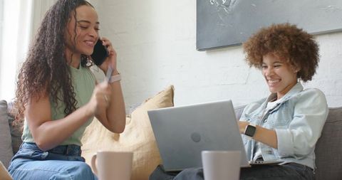 African american friends laughing and working remotely on couch with laptop and phone
