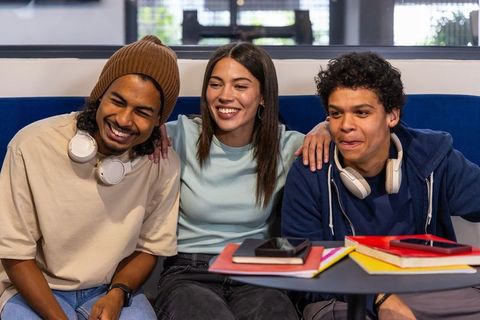 Smiling college friends studying together in campus lounge with notebooks and headphones