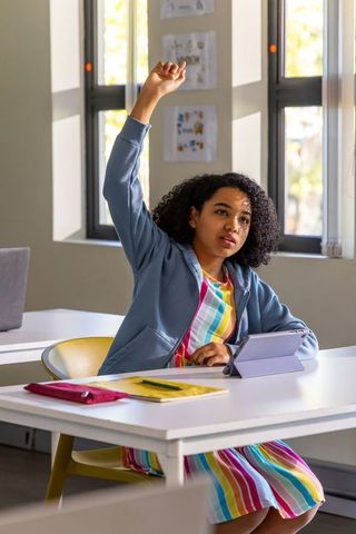 African american teen raising hand while using tablet in bright classroom