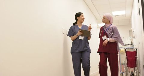 Diverse female nurses walking hospital corridor discussing patient care with tablet