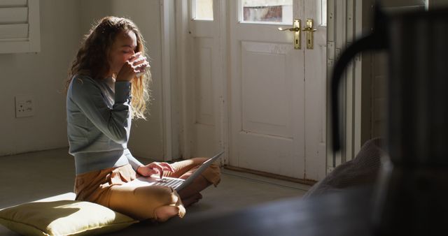 Young woman enjoying a cup of coffee while using a laptop at home. She is sitting on a cushion on the floor by a sunlit doorway, suggesting relaxation and a cozy, homey environment. Ideal for promoting remote work, morning routines, comfortable lifestyles, and home living concepts.