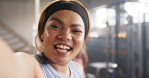 Smiling confident woman taking selfie after workout in gym wearing headband and ribbed tank
