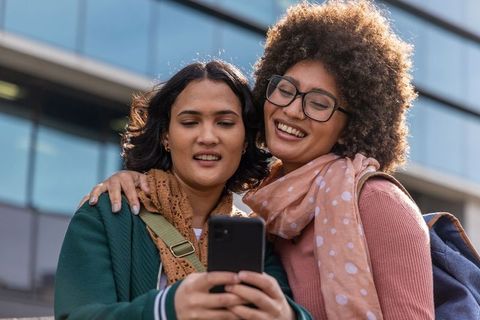 Smiling friends sharing smartphone on college campus with backpacks and scarves