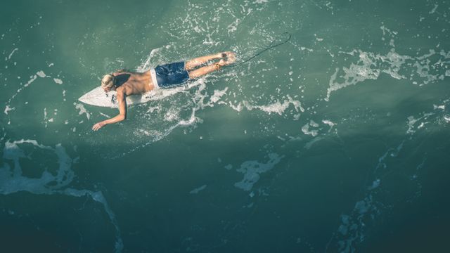Person paddling on surfboard in ocean from aerial view. Surfing lifestyle and summer activity concept. Useful for themes related to surfing, ocean sports, summer vacations, and recreational activities.