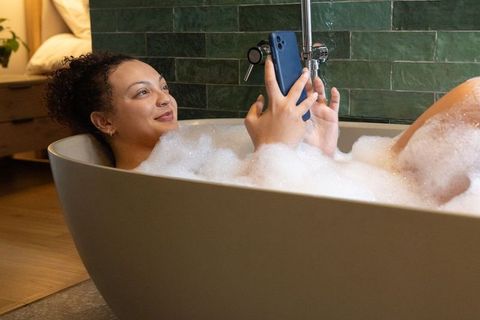 African american woman relaxing in bubble bath using smartphone in modern freestanding tub