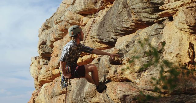 A man is rock climbing outside on a sunny day, wearing proper safety gear like a helmet and harness while secured with ropes on a rugged, rocky wall. This image reflects themes of adventure, outdoor activities, and an active lifestyle, and can be used for marketing in outdoor sports brands, adventure travel promotions, or safety gear advertisements.
