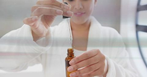 Woman drawing clear facial serum with amber dropper at vanity wearing white robe