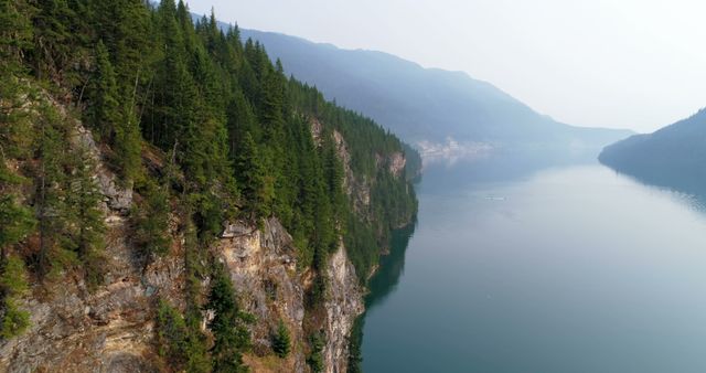A serene landscape showcases a tranquil lake bordered by steep, forested cliffs under a hazy sky. The calm waters and towering trees reflect the untouched beauty of this natural setting.