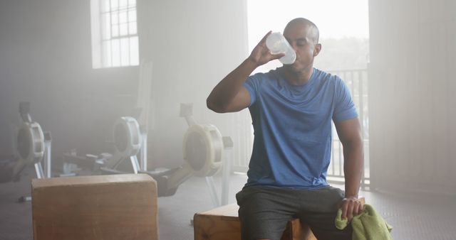 This image shows a man taking a break after his workout in a modern gym. He is drinking water and holding a towel, which implies he is rehydrating and cooling down. The equipment in the background indicates an industrious and active environment. This image is perfect for fitness blogs, health and wellness articles, workout advertisements, hydration campaigns, and gym promotions.