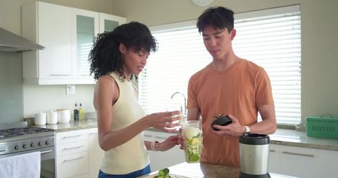Multicultural couple preparing green smoothie with personal blender in bright modern kitchen