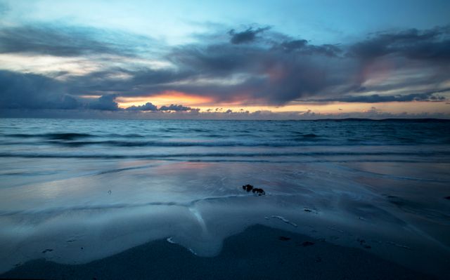 This image shows a tranquil beach scene at twilight, with the sun setting over the horizon. Gentle waves are lapping at the shore while the cloudy sky reflects in the water. Perfect for use in travel blogs, meditation apps, desktop wallpapers, or marketing materials for resorts and vacation destinations due to its relaxing and serene atmosphere.