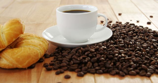 Freshly brewed coffee in white cup on saucer accompanied by croissants and scattered coffee beans on wooden table. Ideal for use in ads and promotions related to breakfast, bakery, coffee shops, and morning routines.