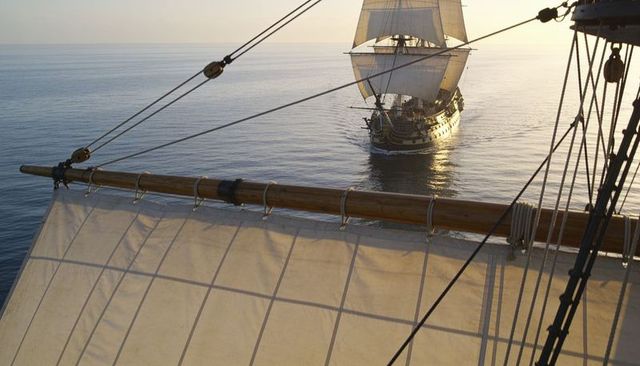 Gliding square-rigged ship reflecting on golden sea behind canvas sail and rigging at dusk