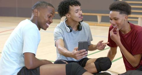 Diverse teen athletes sharing smartphone and laughing while sitting on gym court