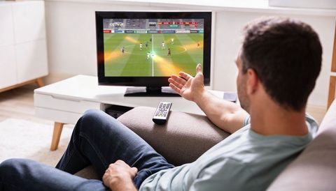 Man watching live soccer match on tv in modern living room with remote on couch arm