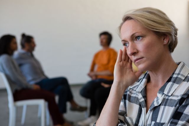 Worried woman sitting with head in hand while friends are discussing in the background. This image can be used to depict themes of contemplation, emotional support, mental health, and group dynamics. Ideal for articles, blogs, or promotional materials related to therapy, support groups, and mental health awareness.