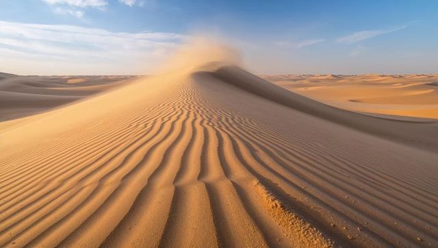 Wind-sculpted golden sand dune ridge with billowing sand plume and rippled desert horizon