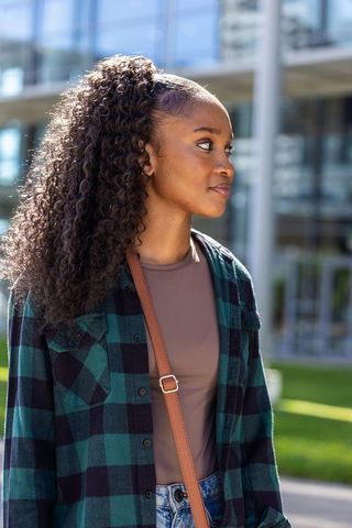 Sunlit campus portrait of young woman in plaid shirt with crossbody bag and modern glass building
