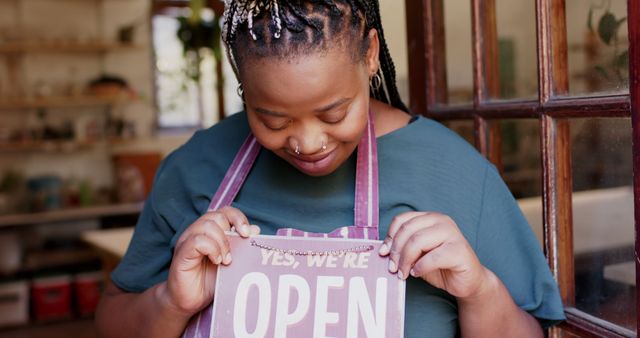 African American female cafe owner smiling and hanging an 'Open' sign at the entrance of the coffee shop. Ideal for topics on small business ownership, entrepreneurship, welcoming environments, hospitality, and local businesses. Use in articles and advertisements promoting small businesses, coffee shops, and community engagement.