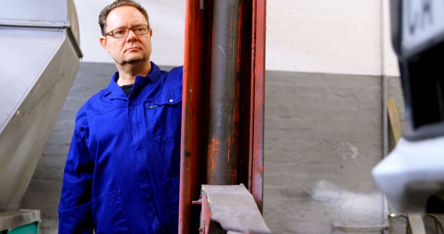 A middle-aged Caucasian man in a blue mechanic's uniform stands thoughtfully in an industrial setting, with copy space. His focused expression suggests he's problem-solving or assessing equipment in a workshop or factory.