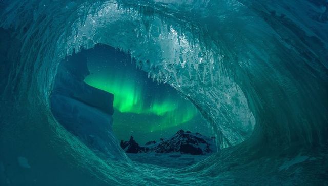 Framing aurora through ice cave arch with icicles and jagged snow mountain silhouettes
