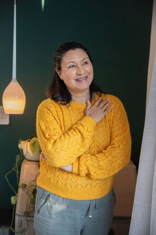 Mature woman wearing mustard sweater holding hand to chest in cozy corner with pendant light