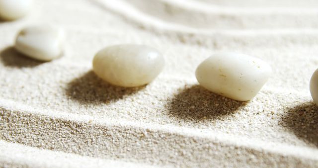 Featuring smooth white stones on sandy Zen garden with raked patterns suggesting tranquility and simplicity. Ideal for themes of mindfulness, meditation, balance, relaxation, and aesthetic presentations. Great for websites, presentations, and promotional materials related to wellness, minimalist designs, and calming environments.