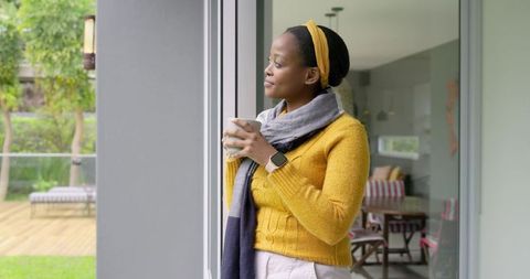 African American woman holding mug at sliding door, looking outside, yellow headband