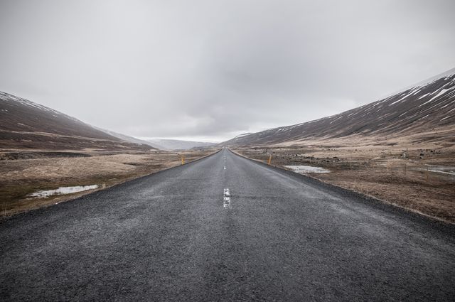 Photograph of a desolate road stretching between barren mountain slopes under an overcast sky. Useful for themes related to travel, adventure, solitude, exploration, and scenic landscapes. Ideal for travel brochures, blogs, wallpapers, and environmental campaigns.