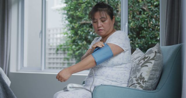 Woman measuring blood pressure using a digital monitor in a cozy living room setting. Suitable for content related to health awareness, self-care routines, healthcare at home, senior care, and wellness lifestyle. Ideal for promoting medical devices, health check-up guides, and content focused on preventive healthcare.