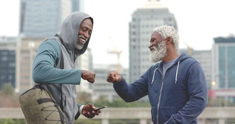 African american senior men bumping fists, smiling friends in hoodies on waterfront