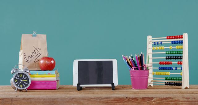 This scene depicts various school supplies neatly arranged on a wooden desk. Items include a digital tablet, a classic alarm clock, a lunch bag, an apple, a stack of books, an abacus, and a container of colorful pencils. Ideal for illustrating educational themes, this image can be used for back-to-school promotions, educational materials, stationery advertisements, or blog posts about school and learning.