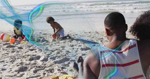 Dad watching kids playing on sunny beach, family relaxing on sand with bright toys by sea