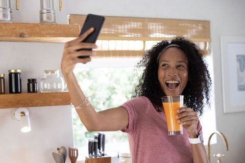 Joyful african american woman taking selfie while drinking orange juice in bright kitchen