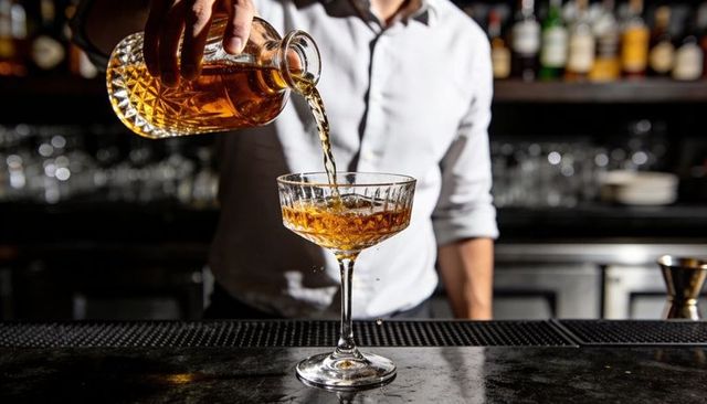 Bartender pouring amber cocktail from cut decanter into crystal coupe glass at bar